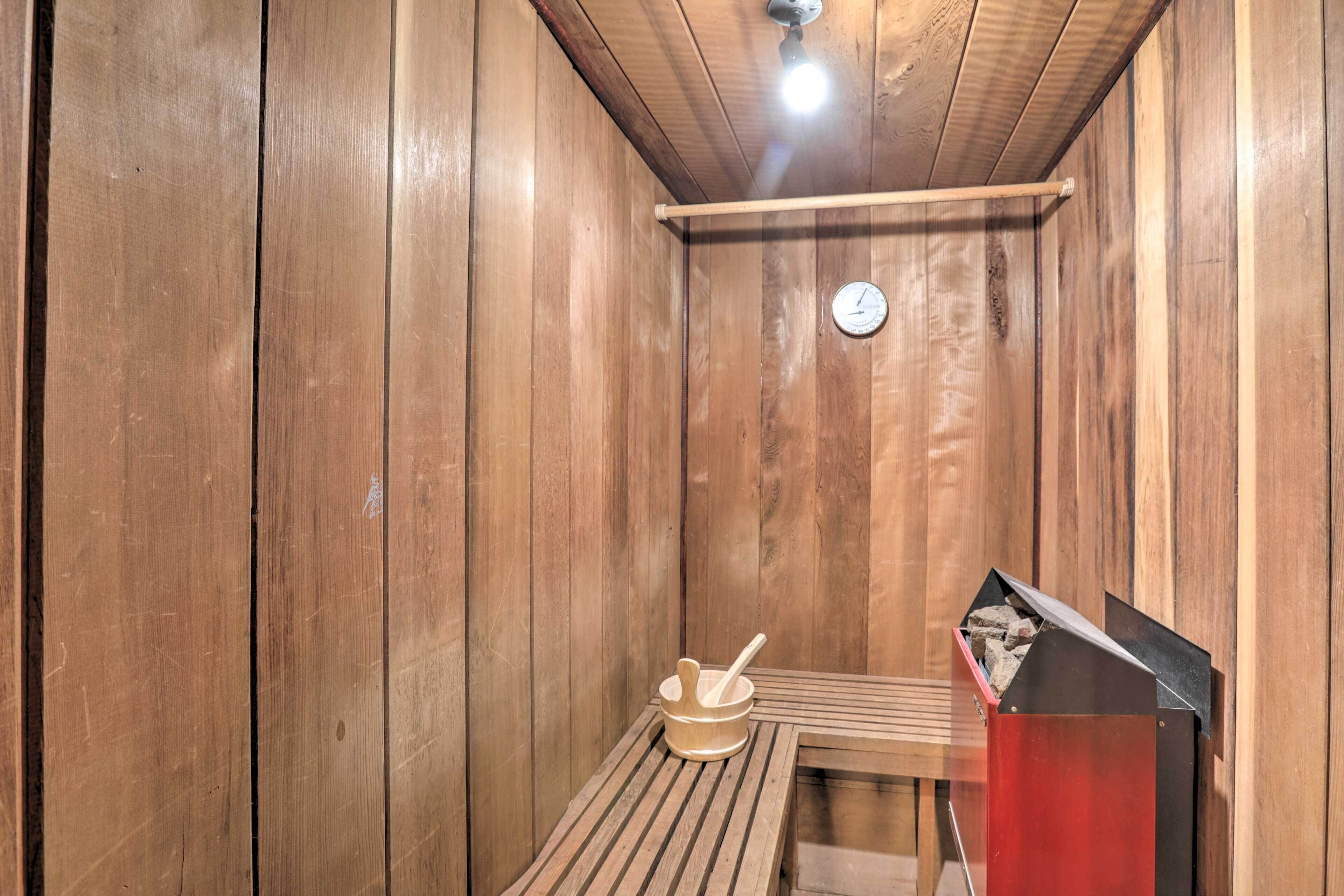 Una sala de sauna con paredes de madera, un banco de madera, un cubo de madera con una cuchara y un calentador de sauna rojo.