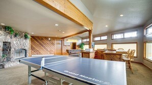 Une table de ping-pong portant l’inscription « Grand Maître » se trouve dans une pièce dotée d’une cheminée en pierre, de poutres en bois et d’un coin repas avec des chaises.