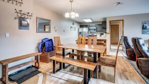Un espace repas avec une table et des bancs en bois, une cuisine à l’arrière-plan et un coin salon avec des fauteuils en cuir.