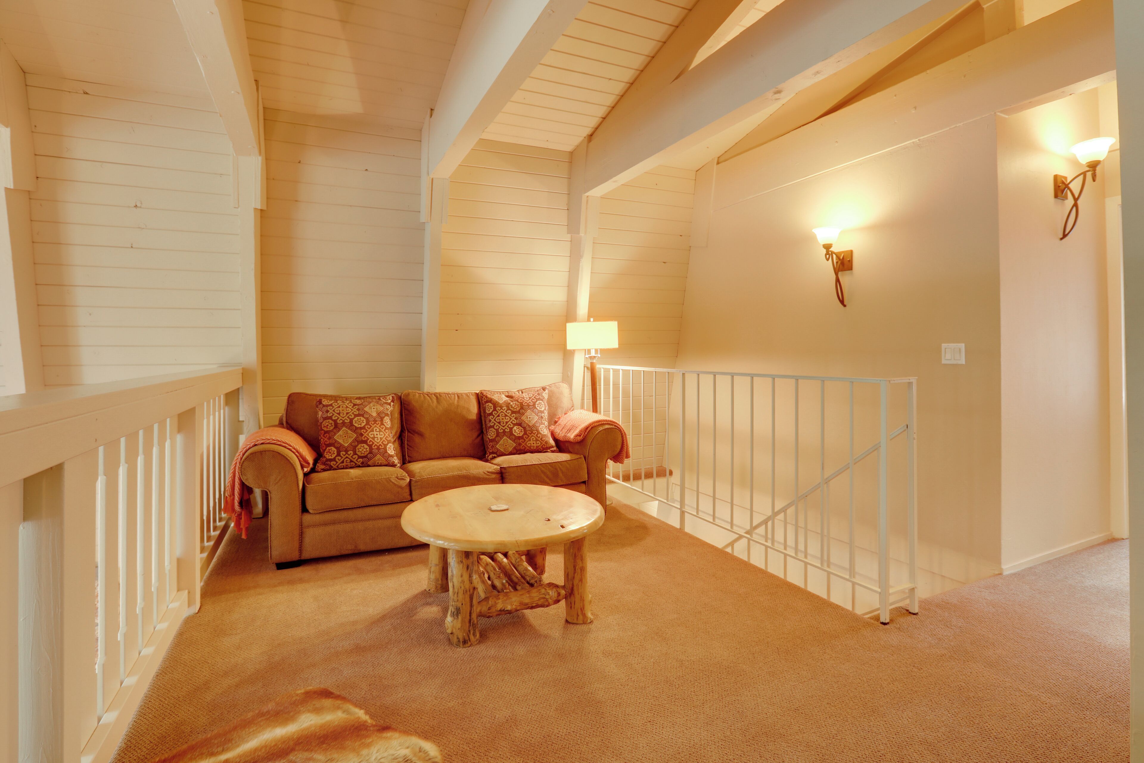 A cozy living area with a wooden ceiling, a beige sofa with patterned cushions, a round wooden coffee table, and a white railing.