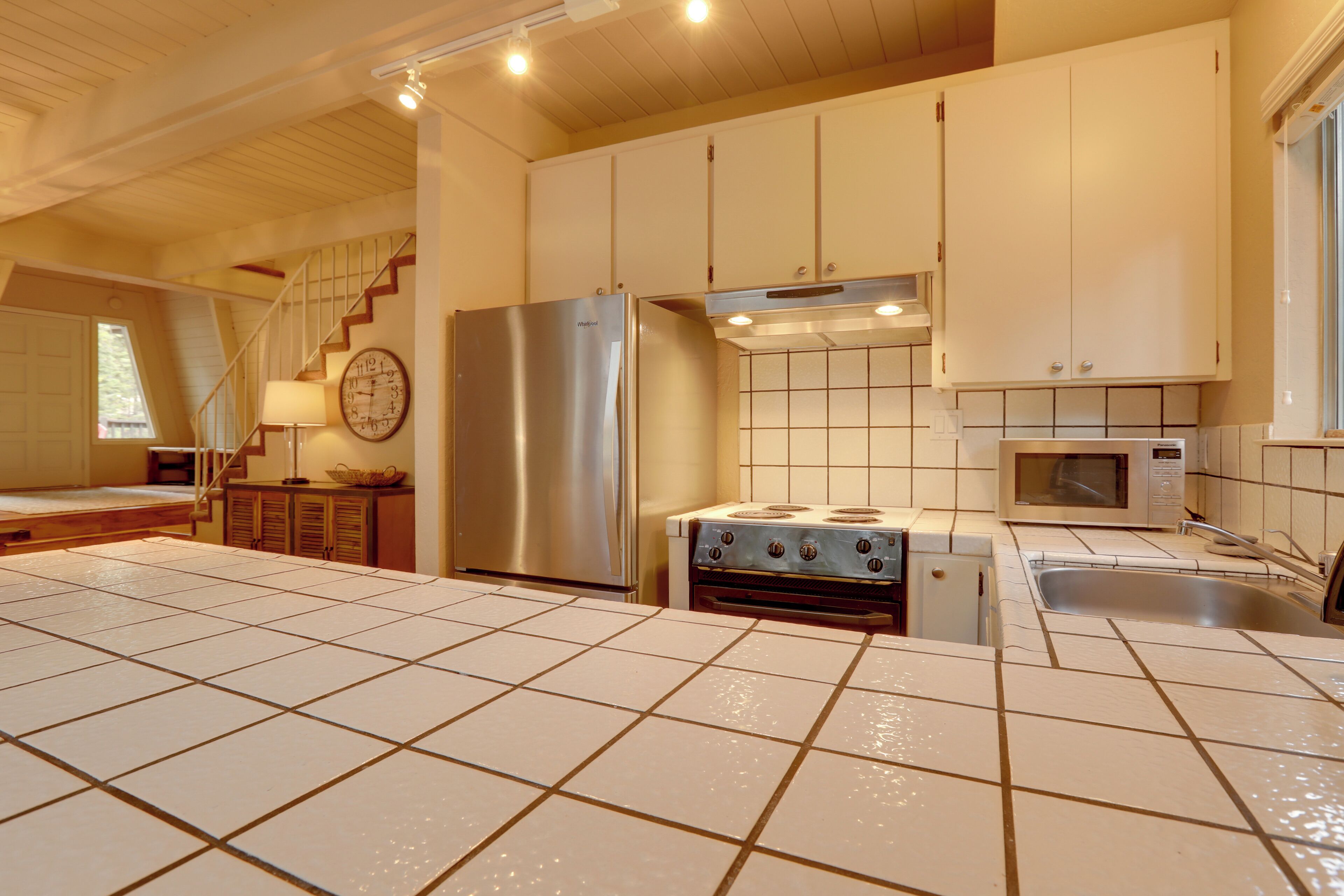A spacious kitchen with a stainless steel refrigerator, a stove, and a microwave. There is a window providing natural light and a clock on the wall.