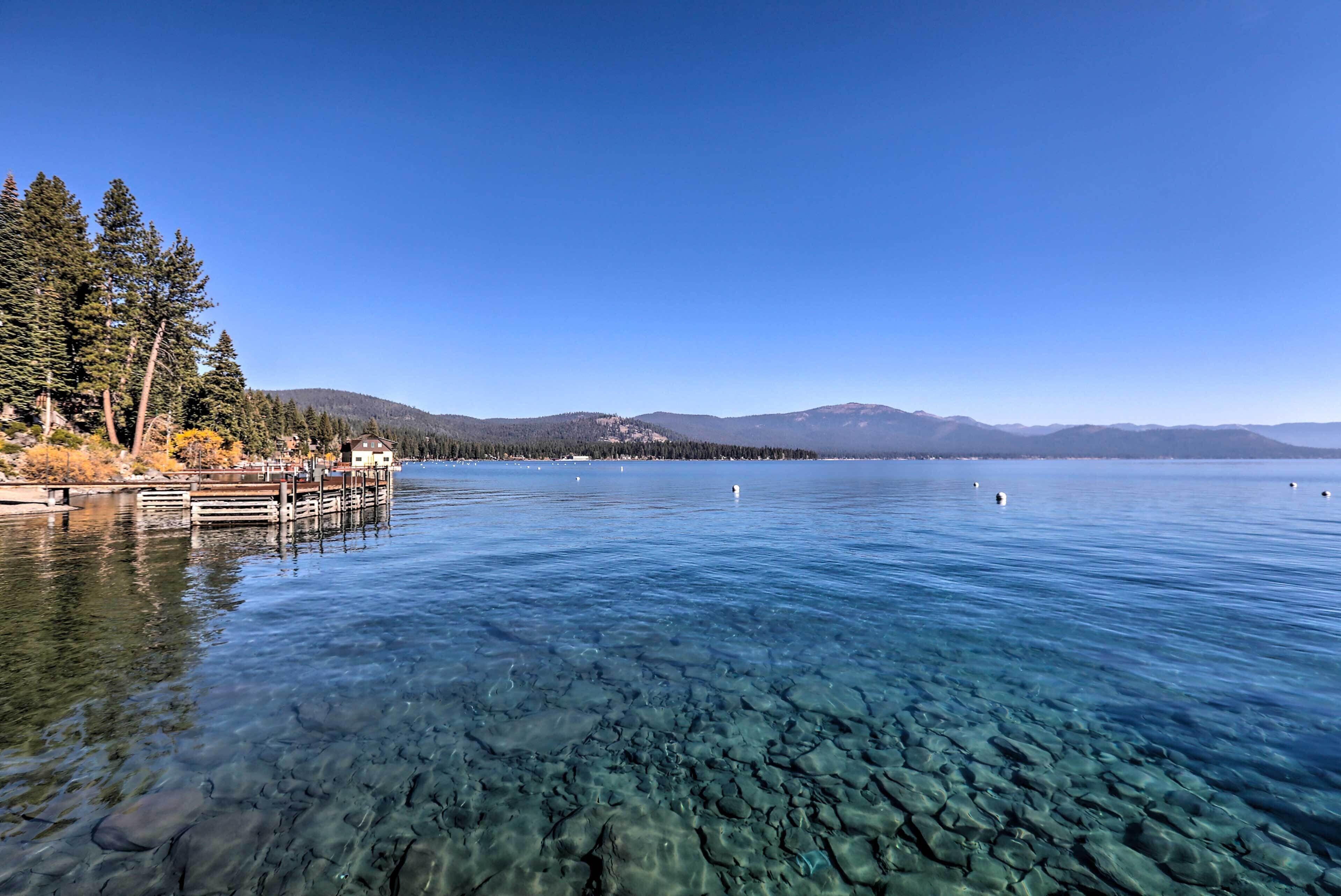 A clear lake with a pier extending into the water, surrounded by trees and mountains in the background.