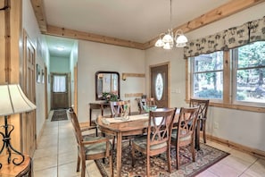 Un comedor con mesa y sillas de madera, una araña de techo y ventanas grandes con vistas a árboles.
