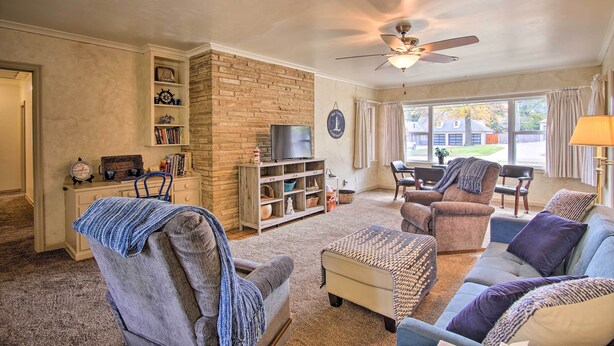 A living room with a stone fireplace, a ceiling fan, and a large window with curtains. - Ranch-style Cottage - 2 Miles to Philbrook Museum! (Tulsa)