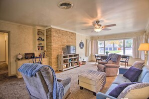 A living room with a stone fireplace, a ceiling fan, and a large window with curtains. - Ranch-style Cottage - 2 Miles to Philbrook Museum! (Tulsa)