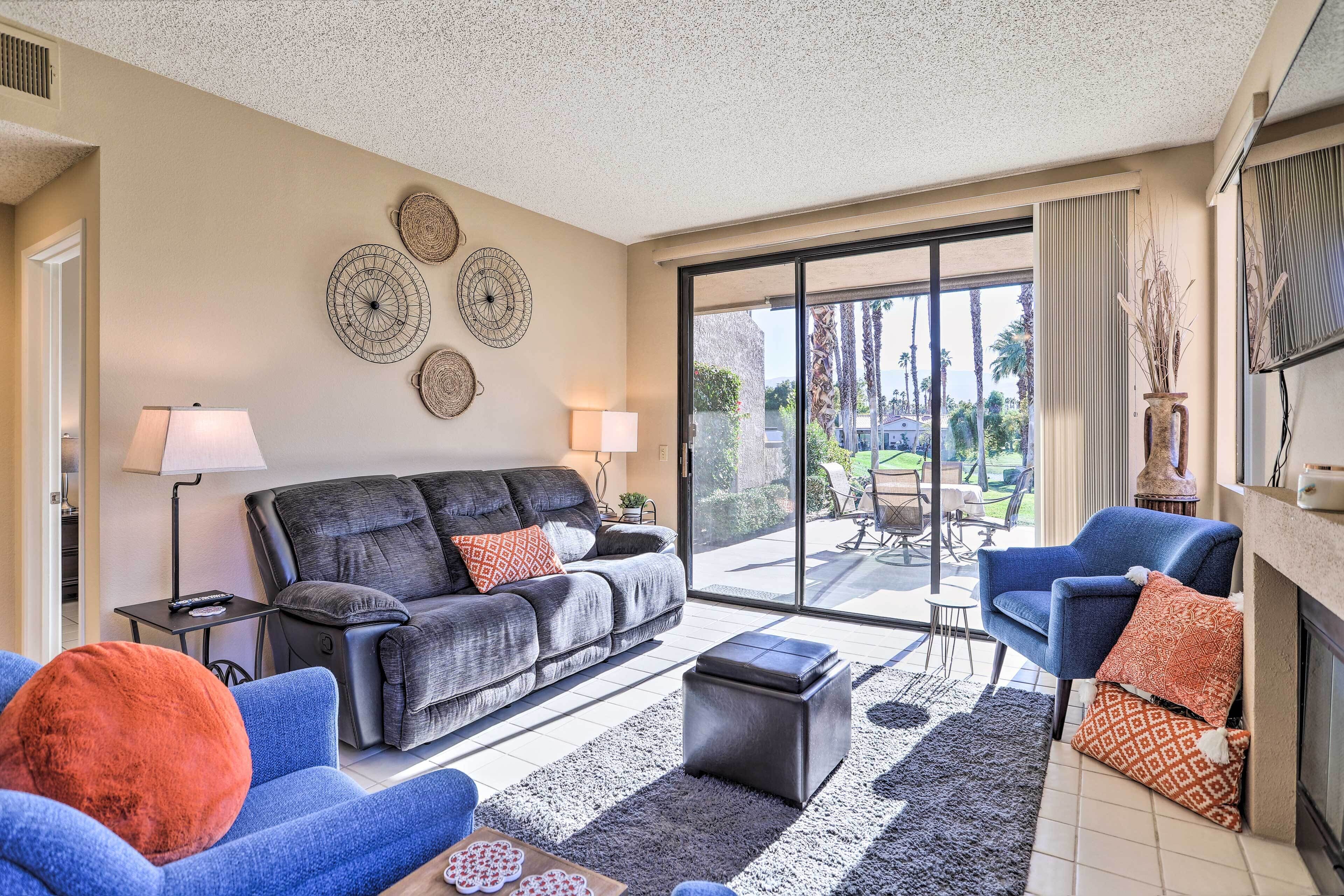 A living room with a sofa, armchairs, a coffee table, and a fireplace. There are decorative wall art pieces and a view through the glass door.