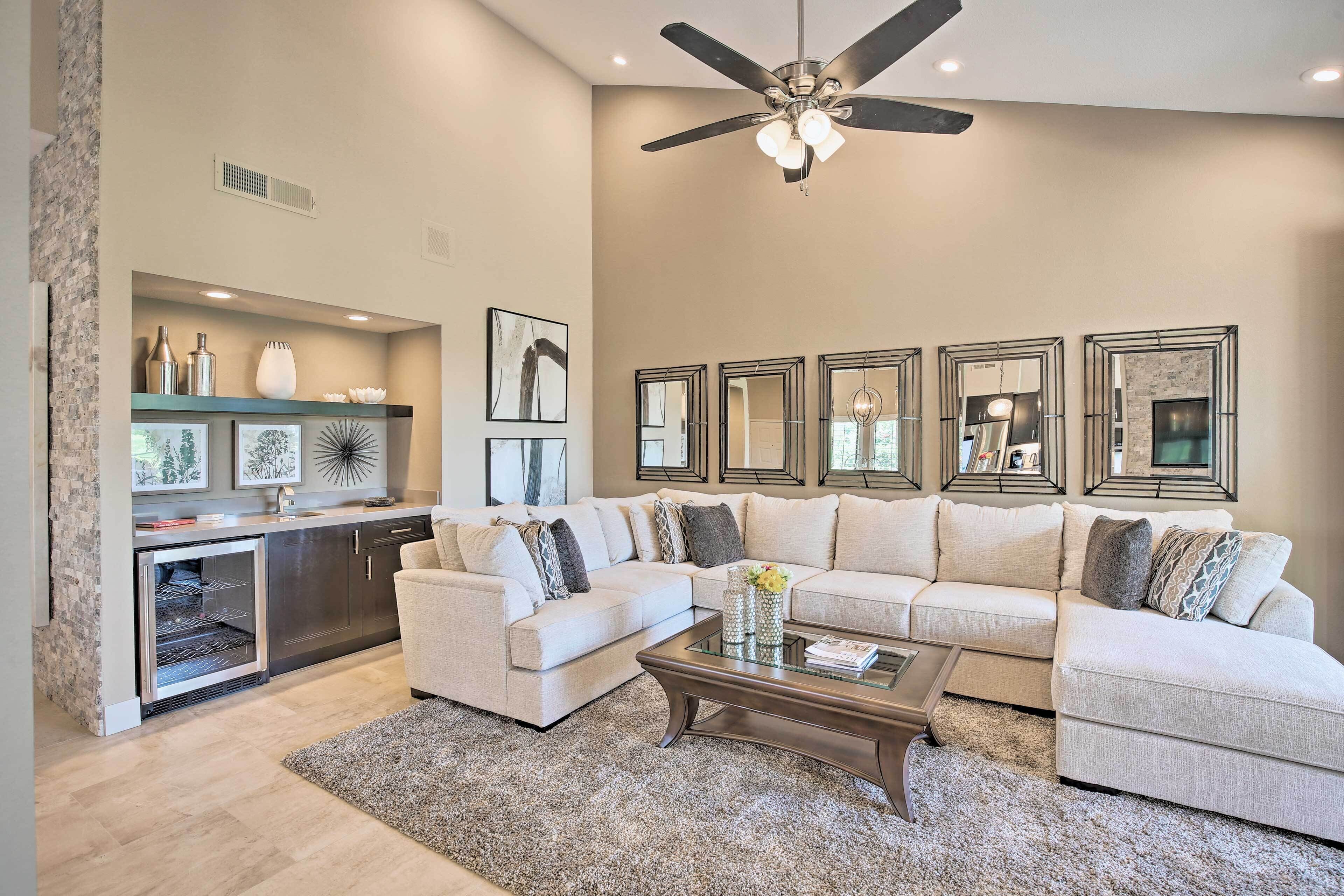 A modern living room with a sectional sofa, a coffee table, and a built-in kitchen area with a refrigerator and shelves.