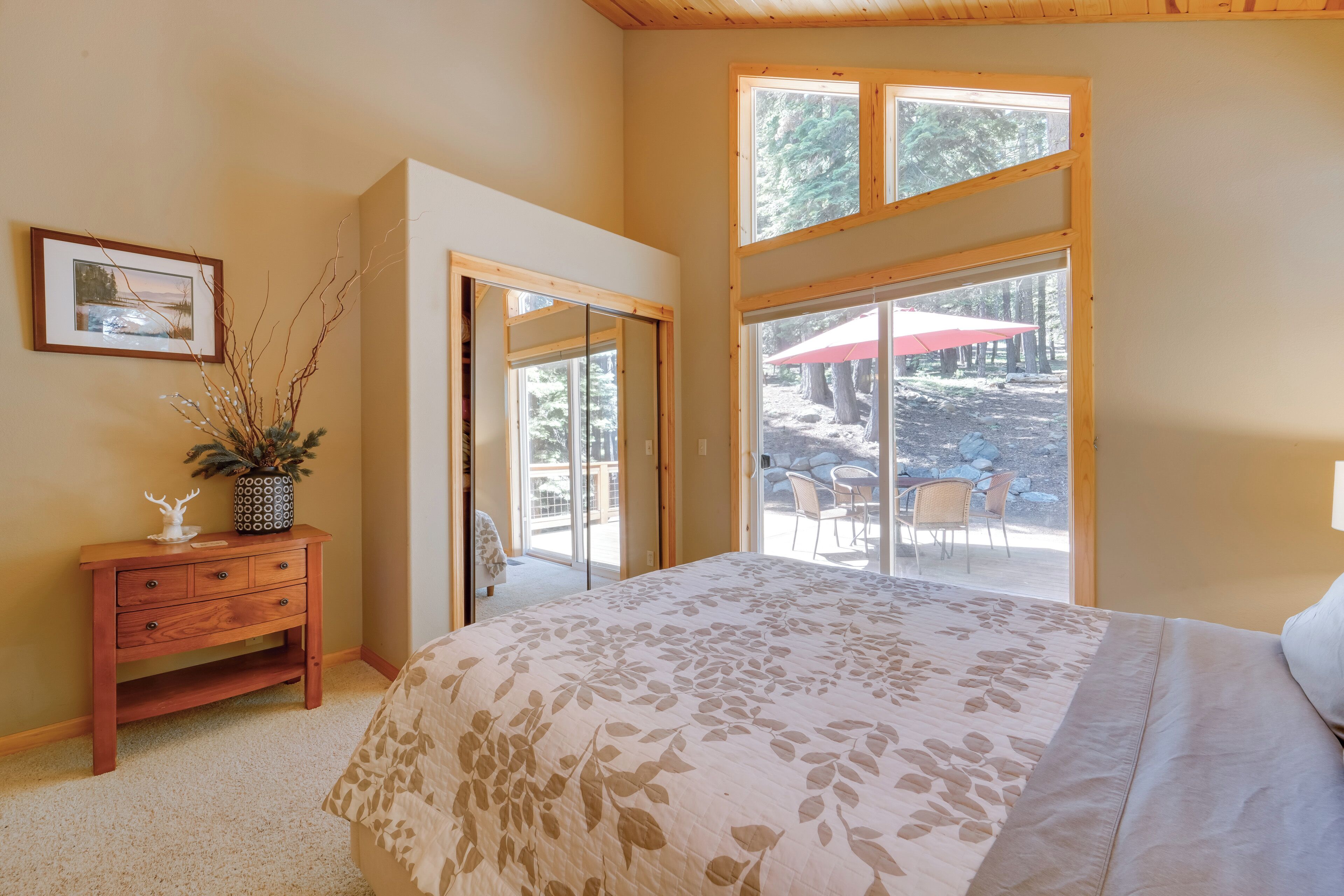 Un dormitorio con una cama grande, una mesita de noche de madera, un cuadro enmarcado y una ventana con vista a un patio con mobiliario exterior.