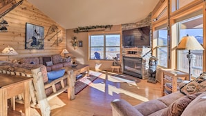A cozy cabin interior with a wooden log wall, a fireplace, a large window, and a sofa.