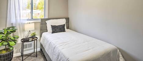 Ein Schlafzimmer mit einem Einzelbett, einem Nachttisch mit Lampe, einer Zimmerpflanze und einem Fenster mit Vorhängen.