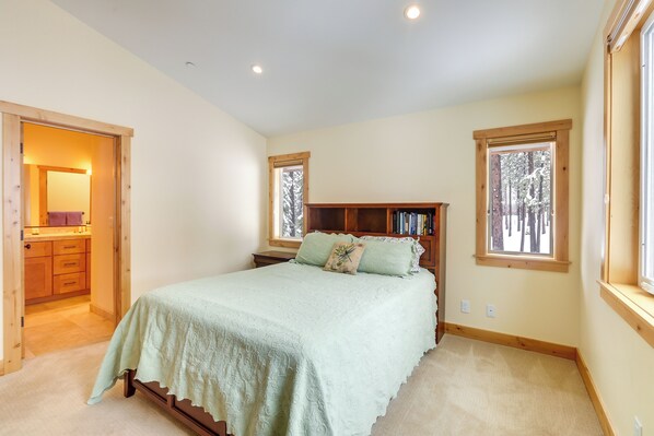 Une chambre à coucher comprenant un lit, une tête de lit en bois, une table de chevet, une bibliothèque et deux fenêtres donnant sur des arbres.