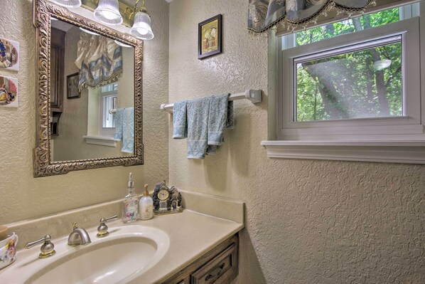 Un baño con un espejo grande y decorativo, un lavamanos con dos grifos y una ventana con vistas a árboles.