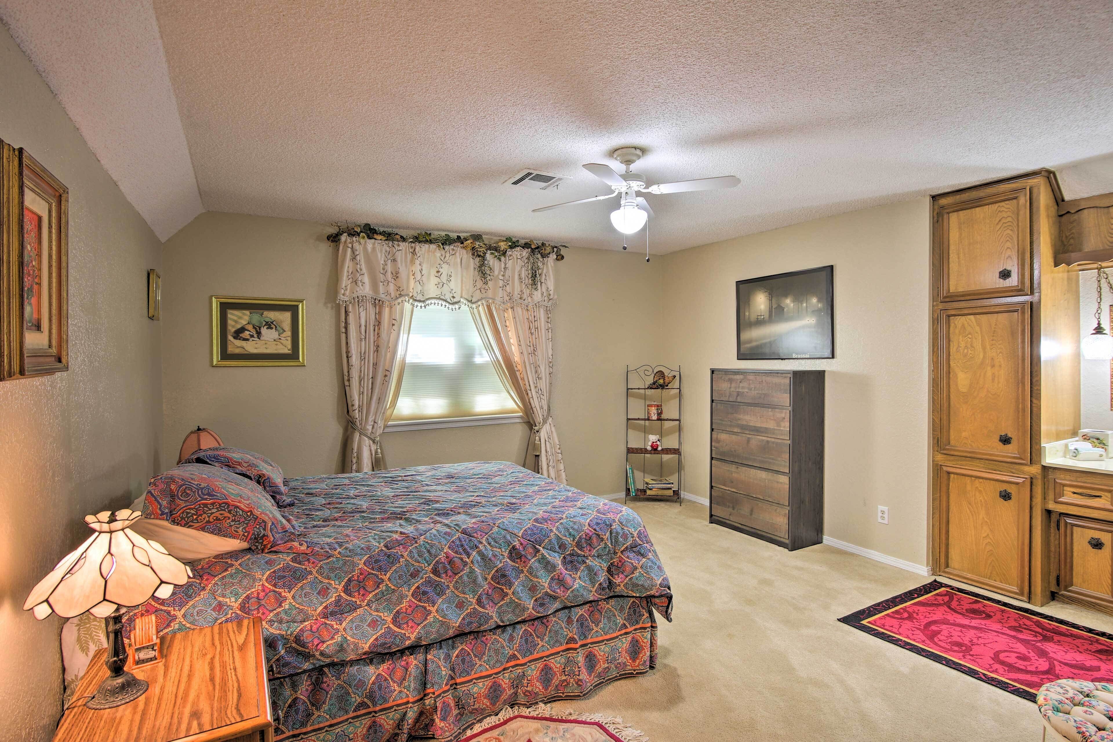 A bedroom with a bed, a ceiling fan, a window with curtains, a bedside lamp, a dresser, and a rug.