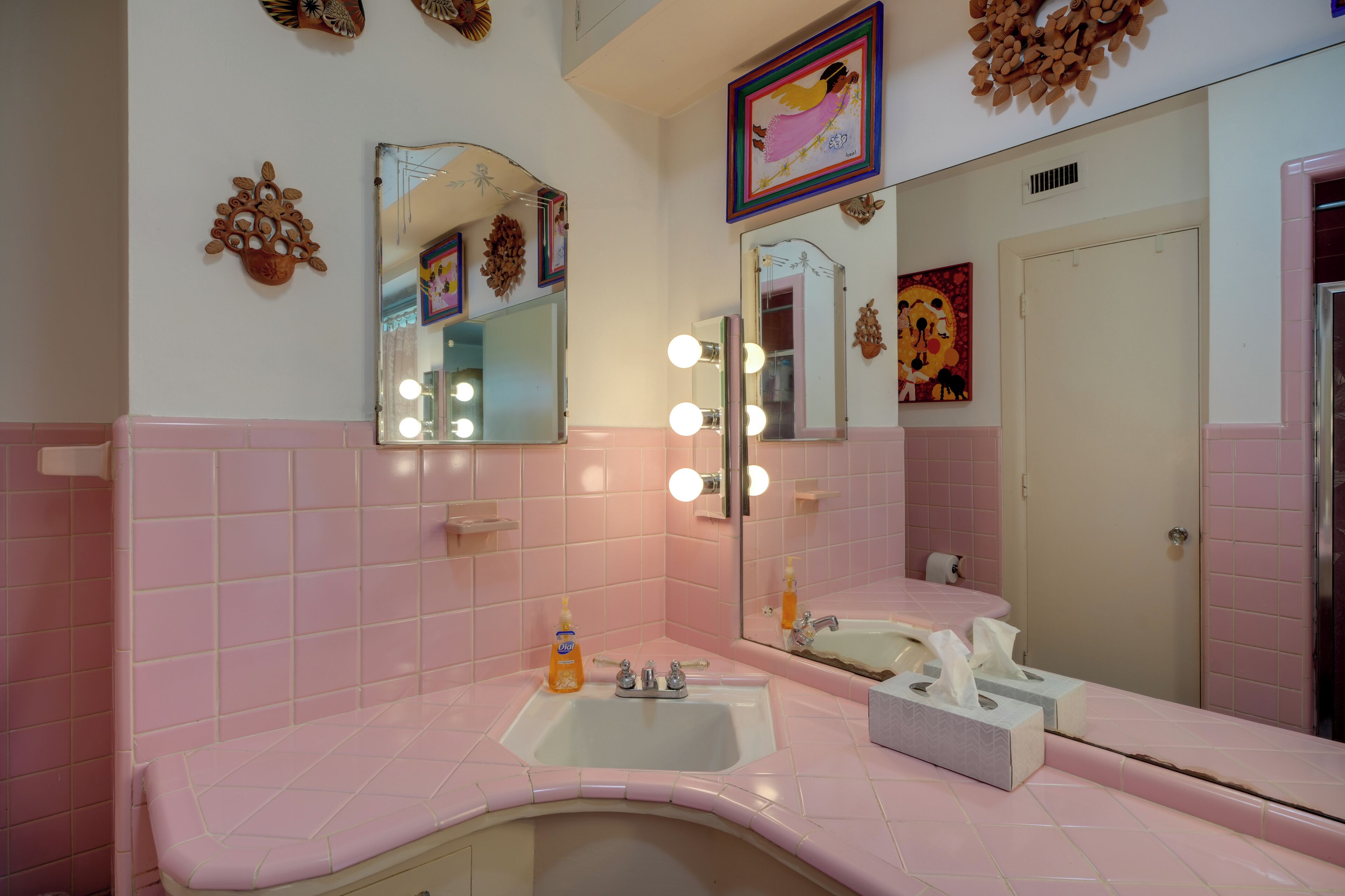 A bathroom with pink tiles, a sink, a mirror with lights, and decorative wall art.