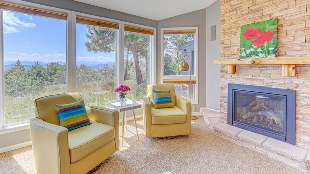 Una habitación con chimenea, dos sillones, una mesita con un florero y ventanas amplias que ofrecen vistas a árboles y montañas.