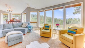 Amplia sala de estar con un sofá modular, dos sillones y un felpudo blanco. Amplias ventanas ofrecen vistas a una zona boscosa.