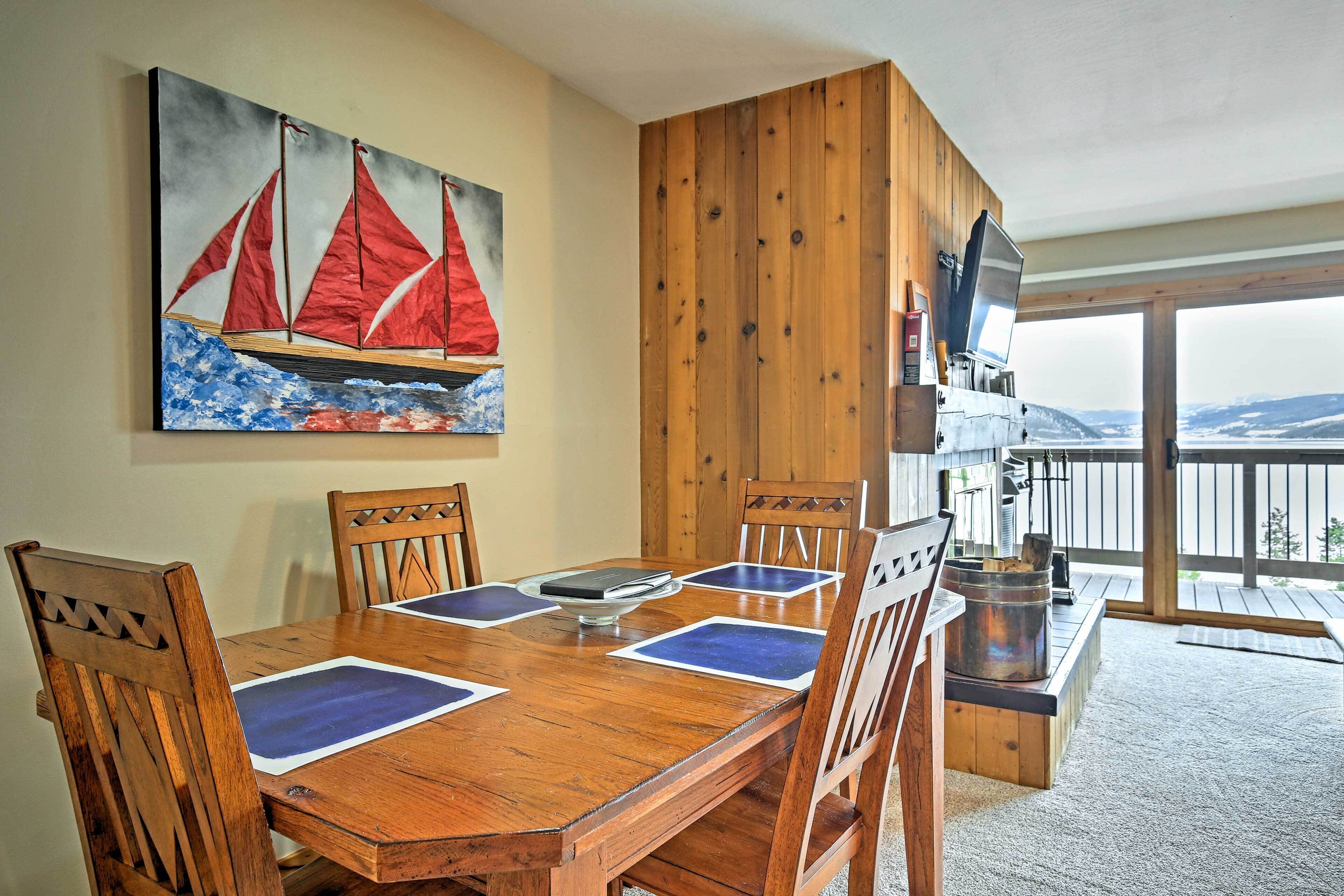A wooden dining table with blue placemats, surrounded by wooden chairs. A painting of sailboats is on the wall, and a television is mounted on the wall.