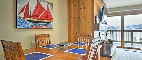 A wooden dining table with blue placemats, surrounded by wooden chairs. A painting of sailboats is on the wall, and a television is mounted on the wall.