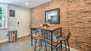 A room with a brick wall, a wooden table with two chairs, a small trash bin, and a window.