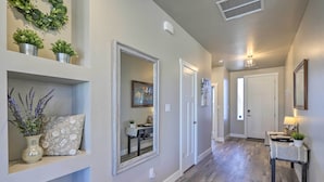 A hallway with a wooden floor, a mirror, and a door. There are potted plants on a shelf and a table with a lamp.