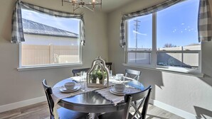A dining area with a round table, chairs, and a chandelier. There are windows with curtains, and a view of a fence and sky.