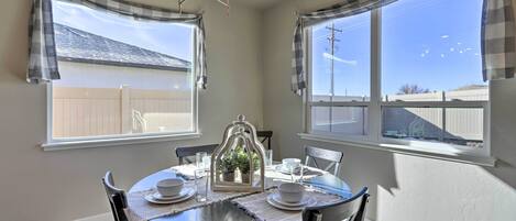 A dining area with a round table, chairs, and a chandelier. There are windows with curtains, and a view of a fence and sky.