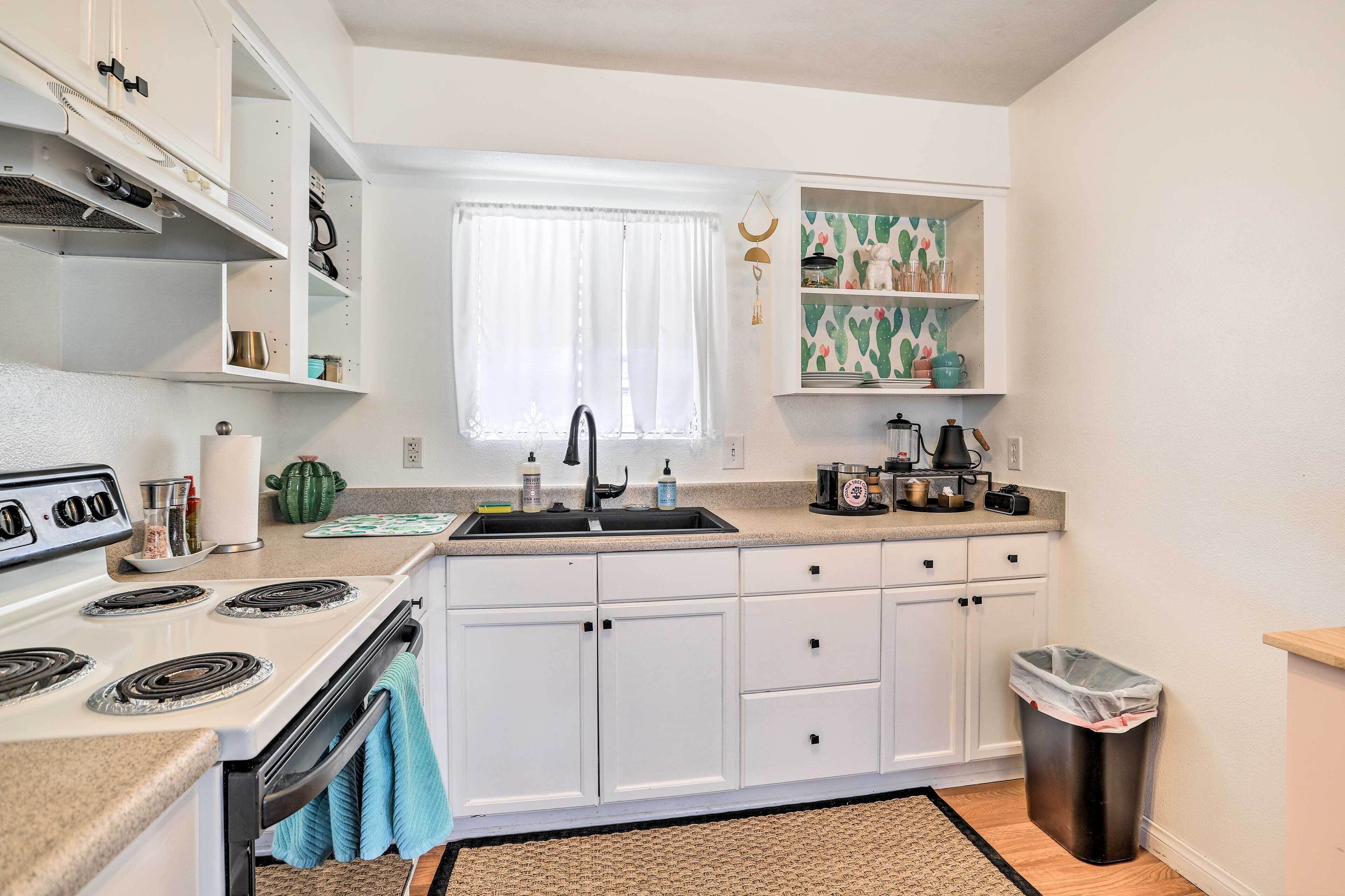 A compact kitchen with white cabinets, a stove, a sink, and a trash bin.