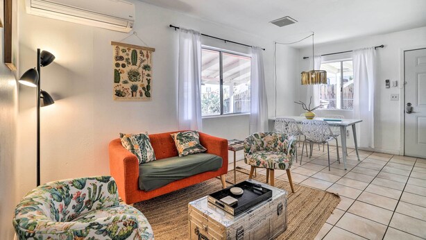A living room with a sofa, armchair, and dining table. There is a window with curtains, a hanging plant, and an air conditioning unit on the wall.