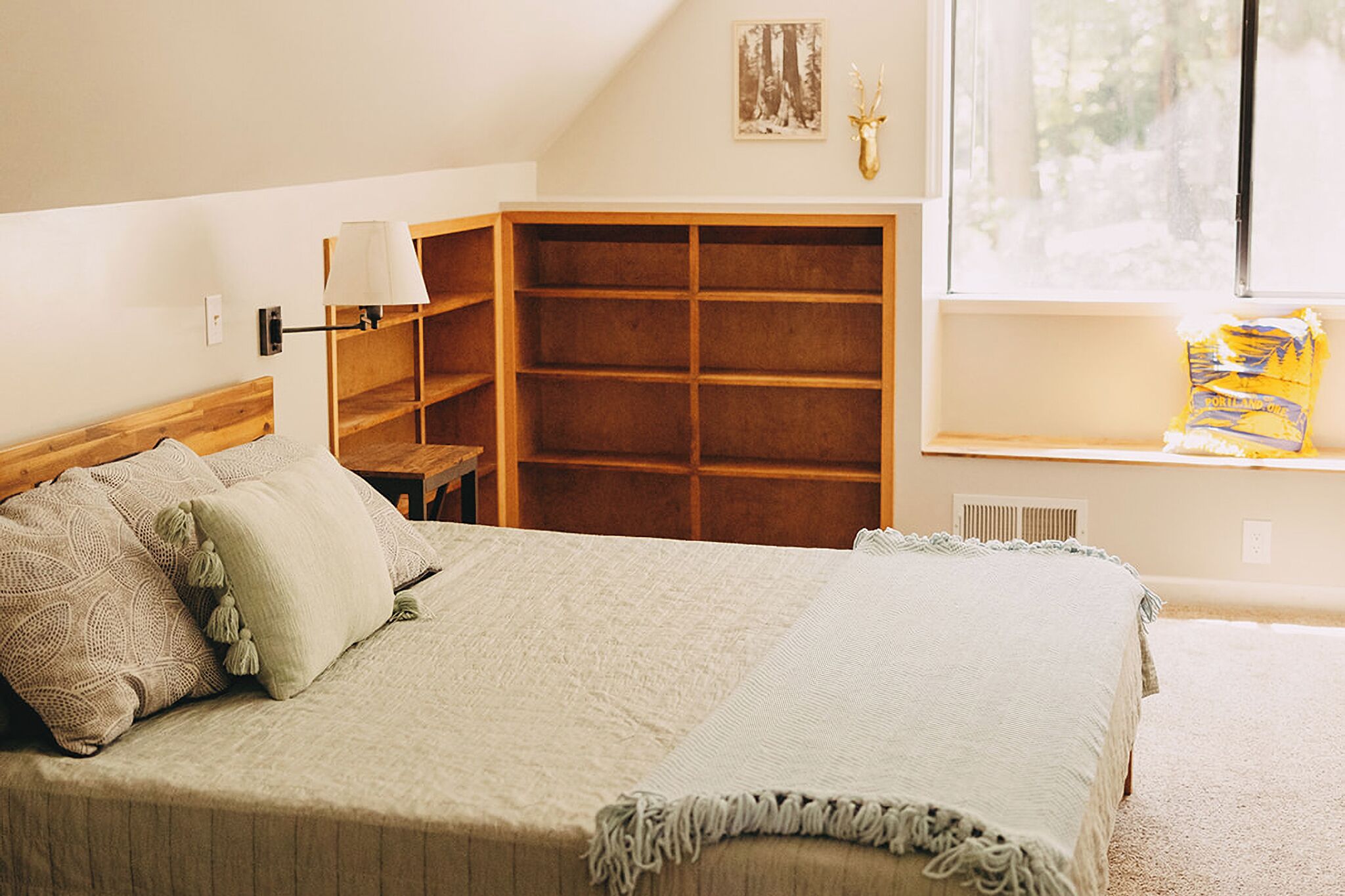 A bedroom with a bed, a wooden shelf unit, a window with a view, and a wall-mounted lamp.