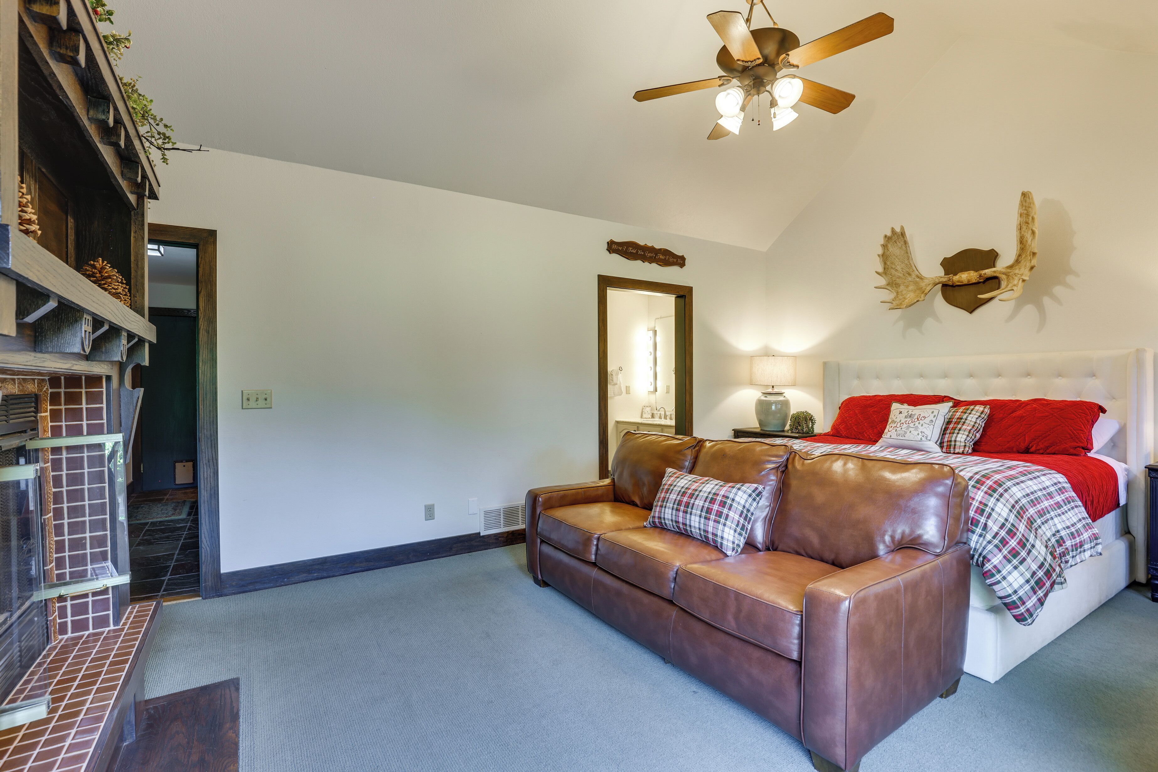 A bedroom with a brown leather sofa, a white bed with red and plaid bedding, a ceiling fan, and a wall-mounted deer antler decoration.
