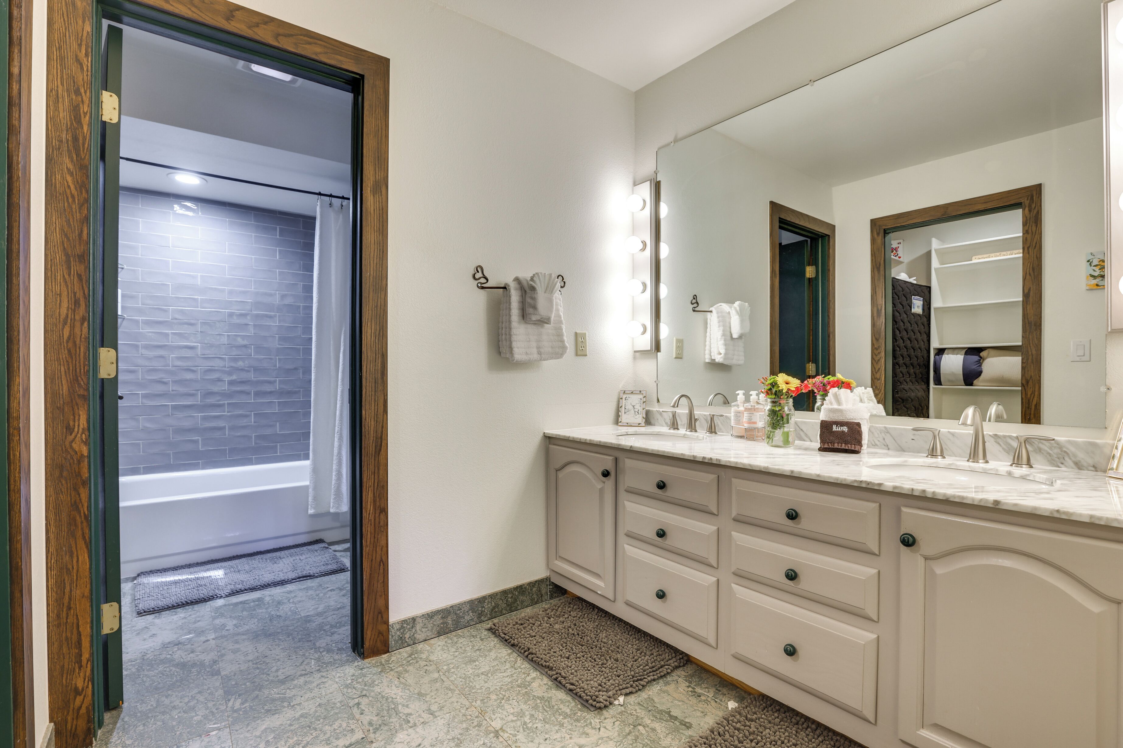 Une salle de bain dotée d’un grand meuble-lavabo, d’un miroir avec éclairage intégré, d’un porte-serviettes et d’une zone de douche visible à travers une porte ouverte.