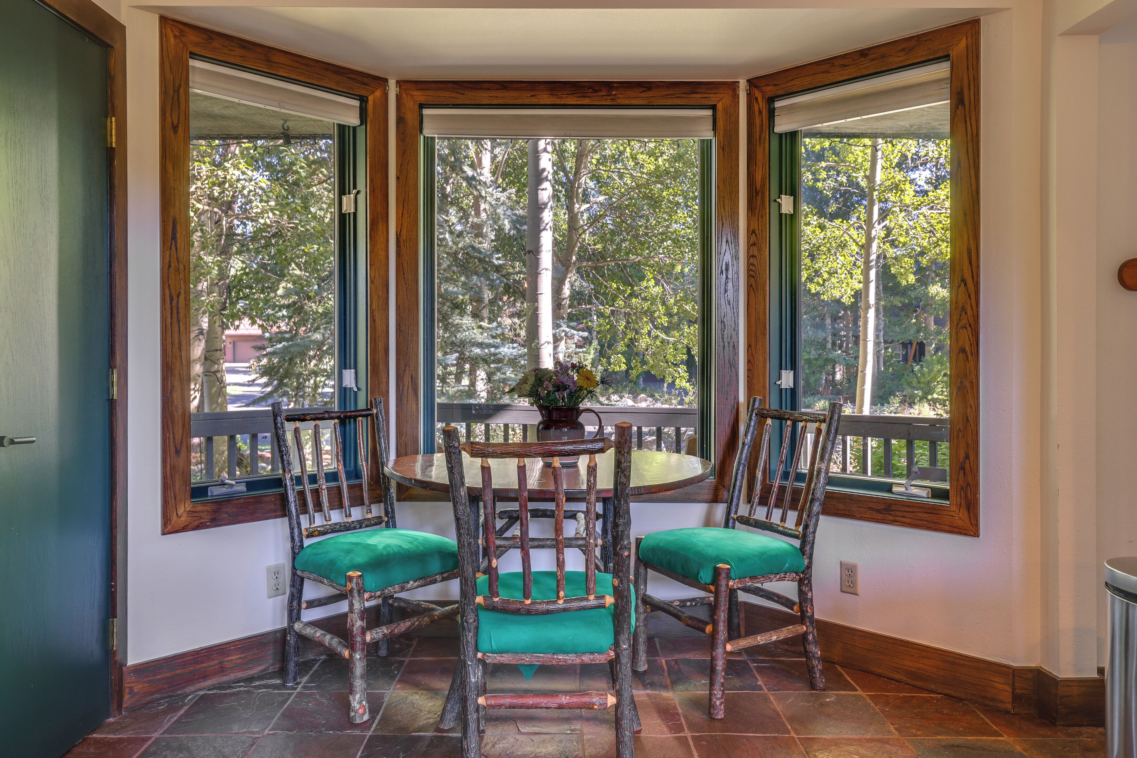 Un comedor con una mesa redonda y cuatro sillas rústicas, ventanas amplias con vista a árboles y un pequeño jarrón con flores sobre la mesa.