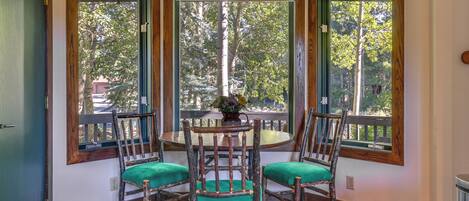 Un comedor con una mesa redonda y cuatro sillas rústicas, ventanas amplias con vista a árboles y un pequeño jarrón con flores sobre la mesa.