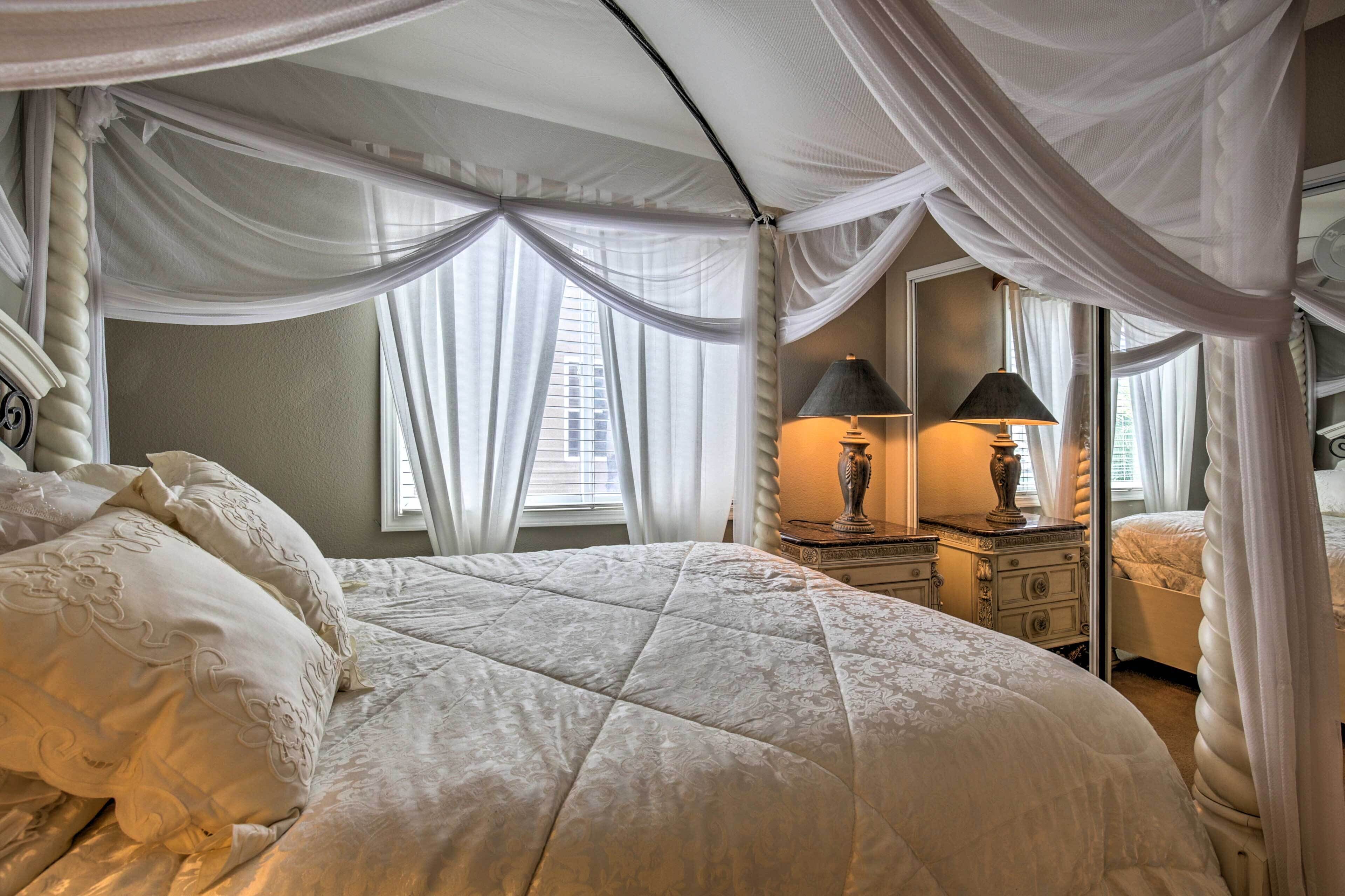 A bedroom with a large bed, a nightstand, a dresser, a mirror, and sheer curtains.