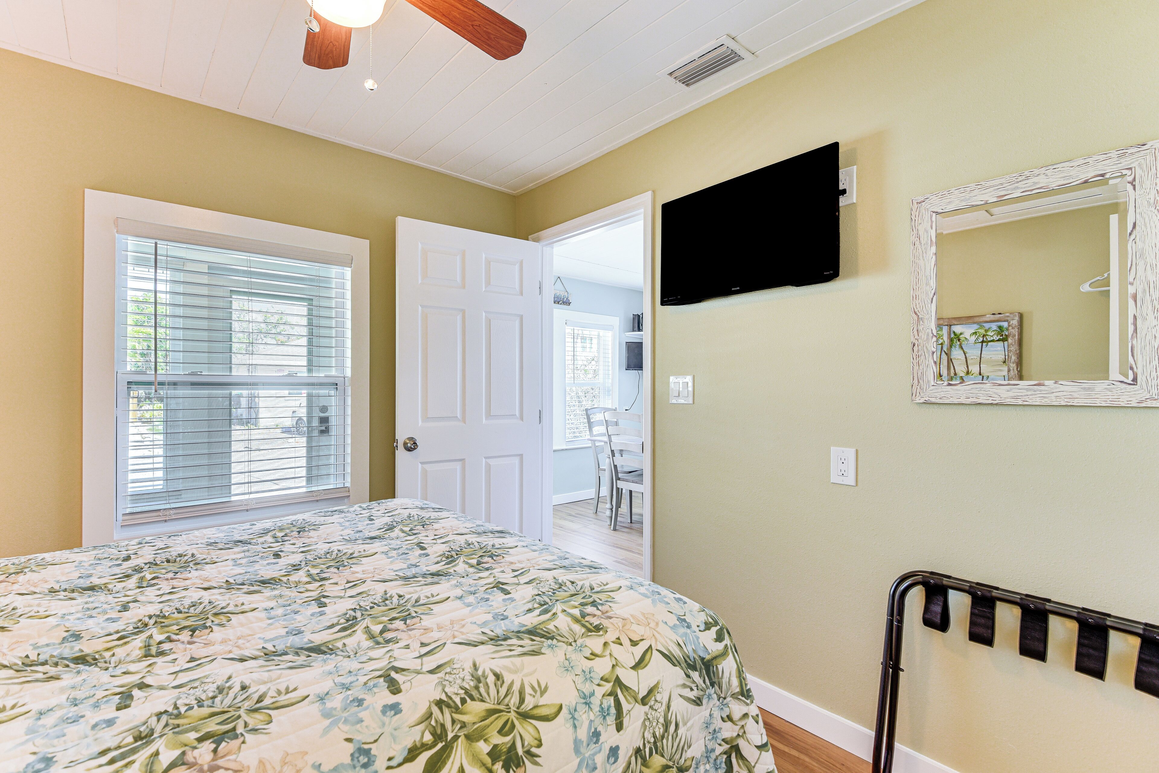 Une chambre à coucher avec une couvre-lit à motifs floraux, un ventilateur de plafond, un téléviseur fixé au mur et une porte donnant sur une autre pièce.