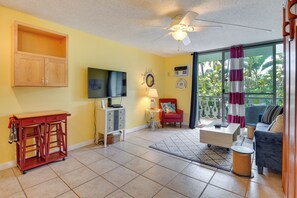 A living room with a TV, a ceiling fan, a red cabinet, a red chair, a blue sofa, and a view of greenery through the window. - Kailua-kona Studio - Walk to Hale Halawai Park! (Kailua-Kona)