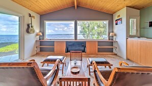 A spacious room with a high wooden ceiling, large windows overlooking the sea, a dining area with wooden furniture, and a kitchen area with wooden cabinets. - Direct Oceanfront, Big Island Home (Keaau)