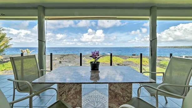 A covered outdoor seating area with a table and chairs, overlooking a beach and ocean. - Direct Oceanfront, Big Island Home (Keaau)