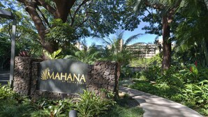 Placa de 'MAHANA at K'Anapali', com um muro de pedra, cercada por vegetação exuberante e um caminho.