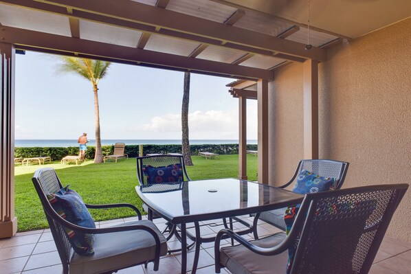 A covered patio with a glass table and chairs, overlooking a lawn and palm tree, with a view of the ocean.
