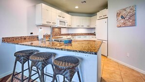 A modern kitchen with white cabinets, a granite countertop, and a built-in microwave. There are three wicker bar stools and a painting of a bird on the wall. - Lanai & Resort-style Perks! Kona Townhome (Kailua-Kona)