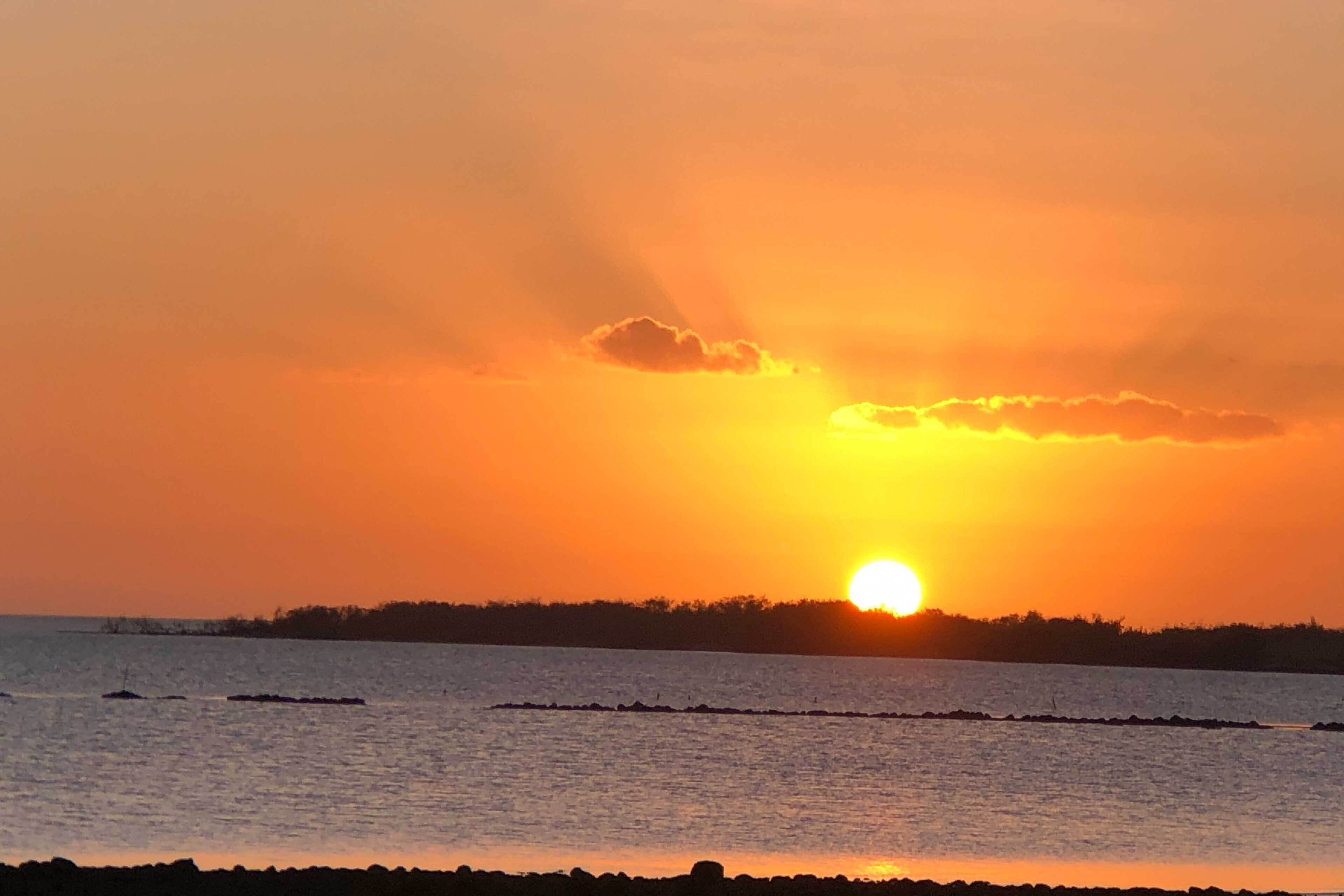 Un coucher de soleil sur une étendue d’eau calme, avec une côte visible au loin.