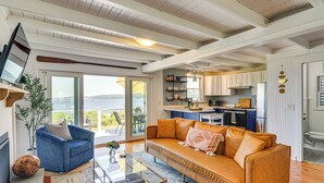A spacious living area with a large sofa, a coffee table, and a television. There is a kitchen area with modern appliances and a view of the outdoors.