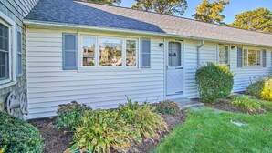 A single-story house with a front porch, a small garden with various plants, and a clear sky. - Romantic Cape Cod Studio w/ Patio, Near Beaches (Dennis Port)