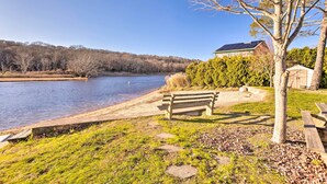 A riverside park with a bench and picnic table, a building with a green roof, and a tree in the foreground. - Cozy Coastal Cottage: 5 Mi to Narragansett Beach! (Narragansett)