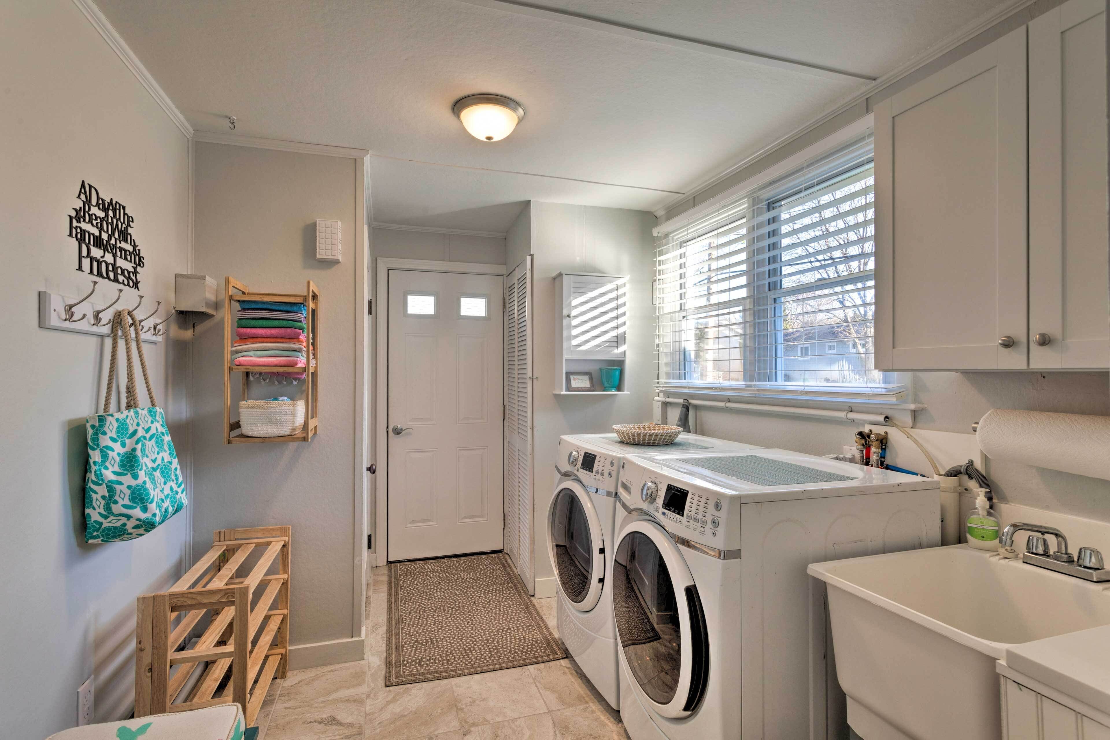 A laundry room with a washing machine, dryer, and sink. There is a door leading to another room, a window with blinds, and a wall-mounted rack with folded towels.