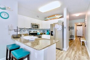 A modern kitchen with white cabinets, a granite countertop, and a built-in microwave. There are two teal bar stools and a small island with a sink. - Beachfront Gulf Shores Condo w/ Pool (Gulf Shores)