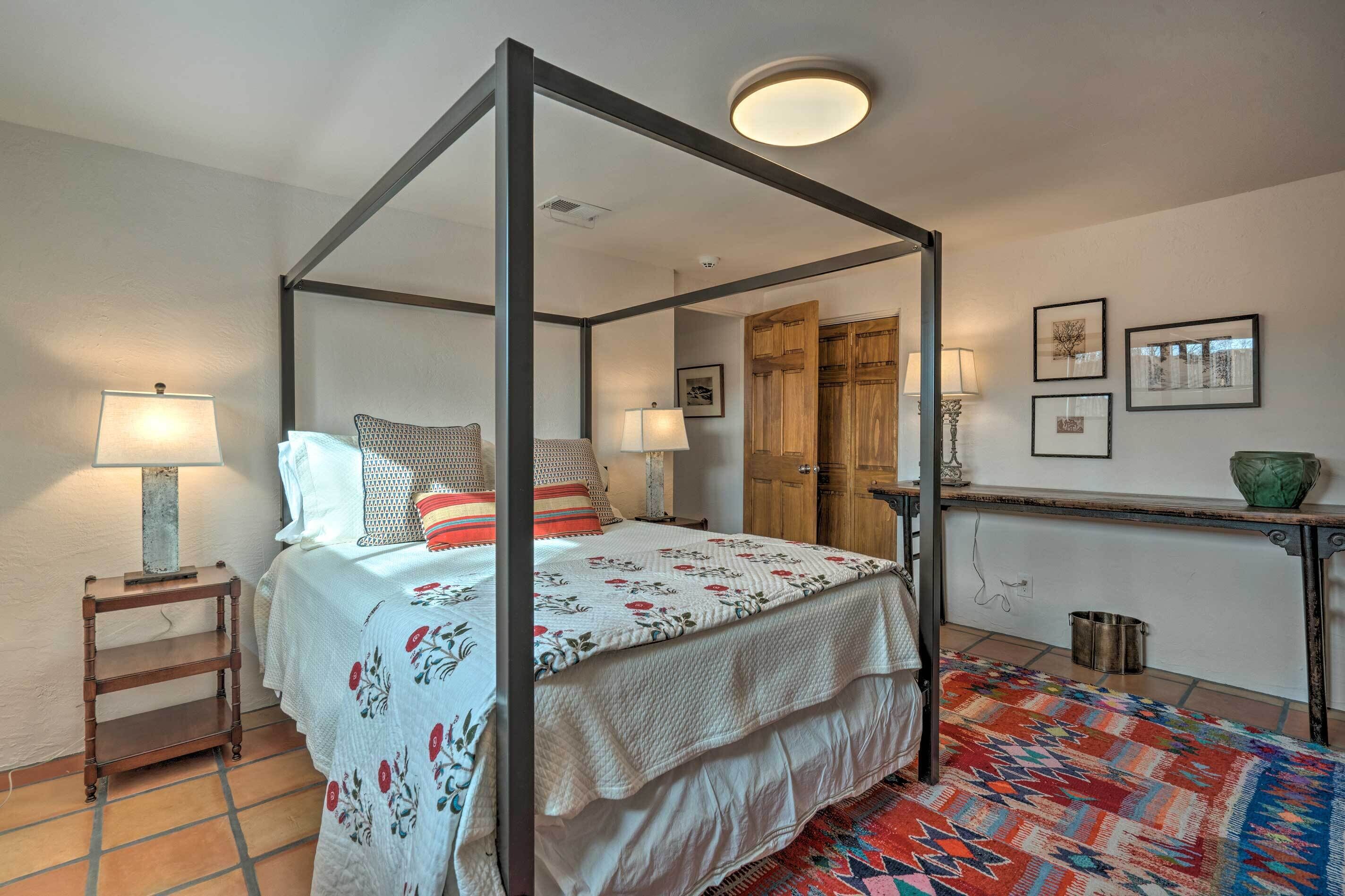 Un lit à baldaquin avec une couvre-lit blanche et des coussins rouges, une table de chevet en bois avec une lampe, un tapis coloré et une petite table avec un bol vert.
