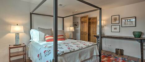 Un lit à baldaquin avec une couvre-lit blanche et des coussins rouges, une table de chevet en bois avec une lampe, un tapis coloré et une petite table avec un bol vert.