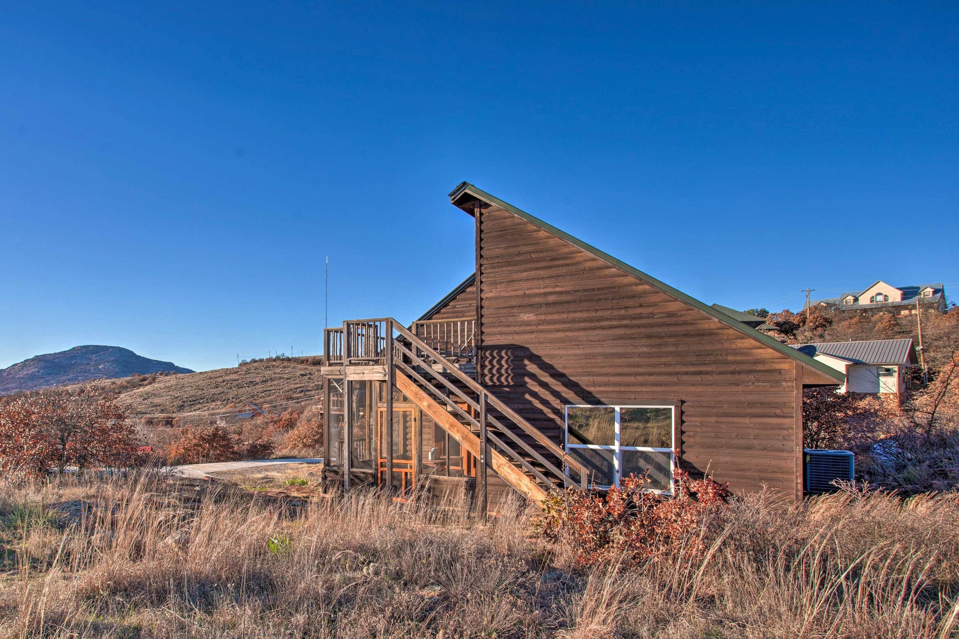 A wooden cabin with a staircase leading to a glass door, surrounded by dry grass and a clear blue sky.